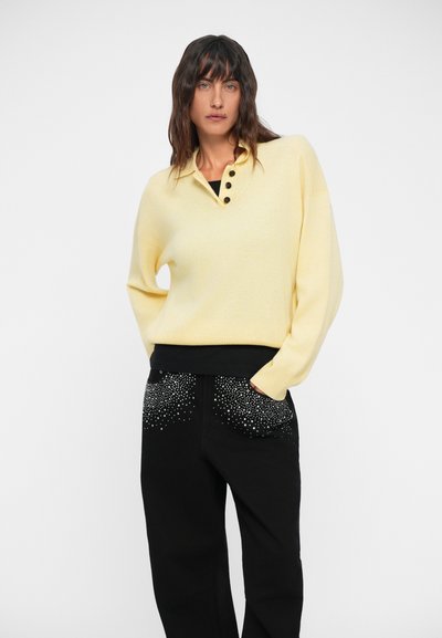 Yellow knit sweater with a collar and three black buttons; paired with black pants featuring studded pocket accents.