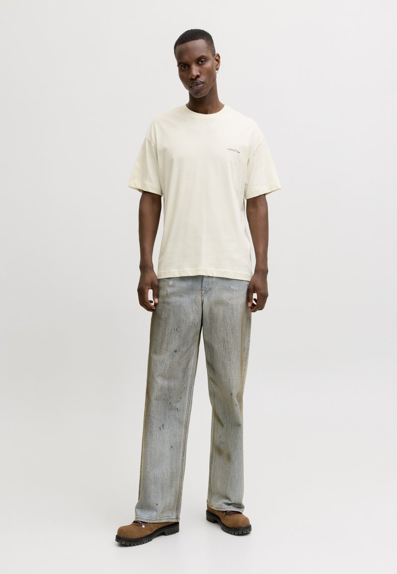 Νέος άνδρας στέκεται φορώντας oversized λευκή μελί T-shirt, χαλαρό ανοιχτό γκρι τζιν με διακριτικούς λεκέδες και καφέ ορειβατικά παπούτσια σε απλό φόντο.