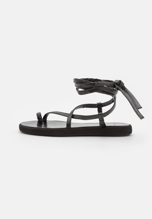 Sandal en cuir noir avec plusieurs fines lanières, un nœud à la cheville et une semelle plate. Texture lisse et design minimaliste.