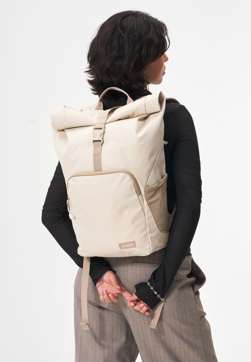 Beige Rolltop-Rucksack mit kontrastierenden Tragen, einer Fronttasche und seitlichen Netztaschen. Hergestellt aus strapazierfähigem Material mit minimalistischem Design.