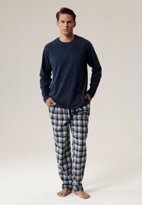 Niebieska koszula z długim rękawem w luźnym kroju i okrągłym dekoltem, zestawiona z piżamowymi spodniami w kratkę w odcieniach granatu, szarości i bieli. Stanie boso.