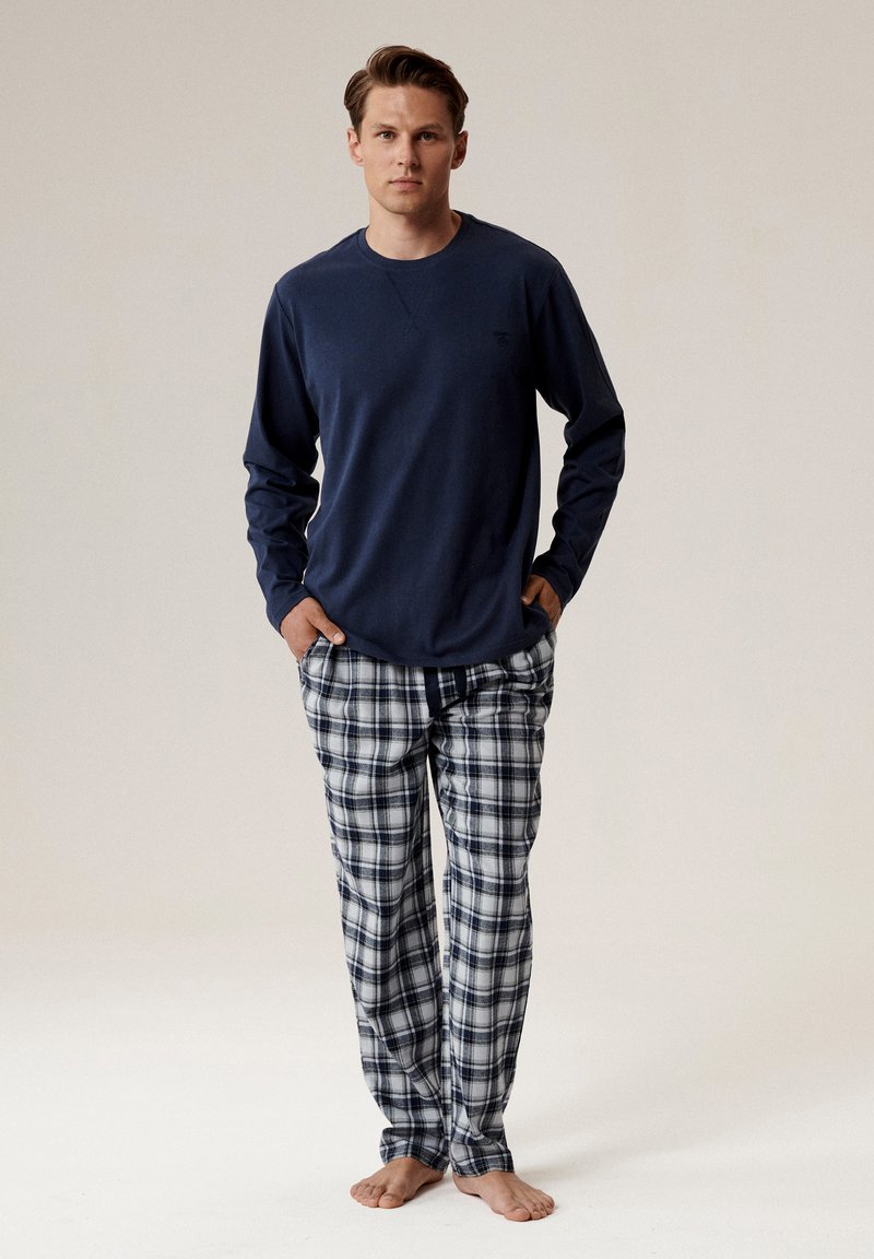 Niebieska koszula z długim rękawem w luźnym kroju i okrągłym dekoltem, zestawiona z piżamowymi spodniami w kratkę w odcieniach granatu, szarości i bieli. Stanie boso.