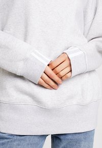 Šedá mikina s volným střihem, žebrovanými manžetami s bílými pruhy a měkkou texturou. Ruce jsou překřížené přes pas.