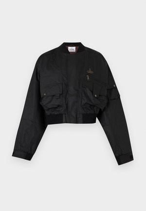 Męski model stoi z ręką w kieszeni, ubrany w czarną kurtkę bomber z wieloma kieszeniami, beżowe spodnie i czarne sneakersy z białą podeszwą.