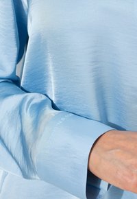 Camicia azzurra a maniche lunghe realizzata in tessuto morbido. Presenta una texture leggermente lucida e coordinate stampate sul polsino.