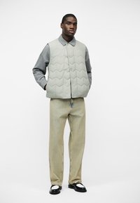 Άνδρας που φοράει ανοιχτό γκρι καπιτονέ γιλέκο πάνω από γκρι πουκάμισο με μακριά μανίκια, μπεζ παντελόνι με φαρδιά μπατζάκια και μαύρα-άσπρα παπούτσια, στέκεται με τα χέρια στις τσέπες.