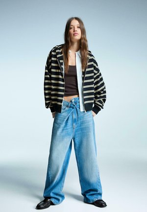 Zwart-wit gestreepte rits hoodie over een bruine tanktop, gecombineerd met lichtblauwe wijdvallende jeans. Model draagt zwarte schoenen.