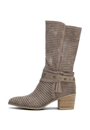 Bota marrón claro perforada hasta la mitad de la pantorrilla con tacón apilado y correa decorativa con tachuelas metálicas y detalle de borla sobre fondo blanco.