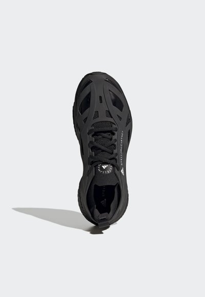 Chaussure de sport noire avec une tige en mesh, des contours façonnés, une semelle épaisse et des accents texturés. Elle dispose de lacets et d'un logo sur le côté.