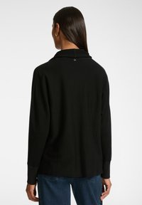 Frau mit langen, dunklen Haaren, die goldene Ohringe trägt, ein schwarzes Langarmshirt und blaue Jeans anhat, mit dem Rücken zu einer einfarbigen Wand gewandt.
