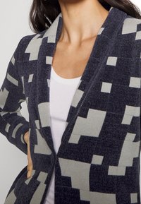 Kvinna som bär en mörkgrå och ljusgrå cardigan med geometriskt mönster över en vit topp med rundad hals, handen vilande på höften.