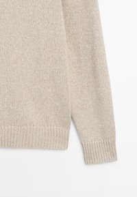 Rukáv a lem béžového pleteného svetru s žebrovanými manžetami, zobrazené na bílém pozadí.