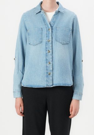 Buttoned light blue denim shirt with two chest pockets worn over a white top, paired with black pants, shown from neck to hips.