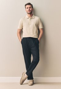 Tricou polo bej, pantaloni negri și mocasini bej. Tricoul are guler și nasturi, iar pantalonii sunt largi. Texturi netede.