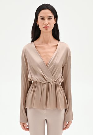 Femme portant un chemisier portefeuille en satin beige avec taille froncée et manches longues, assorti à un pantalon beige assorti sur fond blanc uni.