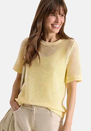 Smilende kvinde med langt brunt hår iført en gul kortærmet mesh-top, beige bukser og med en beige taske på skulderen.