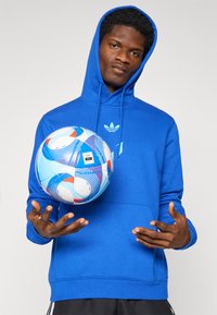 Sweatshirt azul com capuz e bolso canguru, com um logótipo. Uma bola de futebol multicolorida é segurada acima de mãos abertas, exibindo padrões circulares.