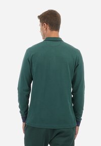 Zelena polo majica s dolgimi rokavi in rebrastim ovratnikom, ki ima kontrastne mornariško modre obrobe na manšetah ter ohlapen kroj.