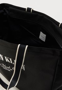 Μαύρη υφασμάτινη τσάντα tote με λευκό λογότυπο, διαθέτει διπλές υφασμάτινες λαβές και εσωτερική τσέπη με φερμουάρ. Ετικέτα ορατή στο εσωτερικό.