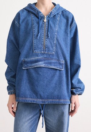 Personne portant une veste à capuche en denim bleu avec fermeture éclair à l'avant, grande poche avant avec rabat, poignets élastiques et pantalon en denim assorti.