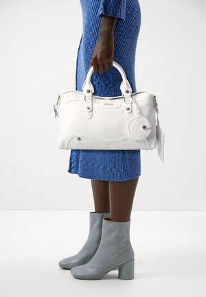 Borsa in pelle bianca con doppio manico, design curvo, hardware argentato e etichetta staccabile. Indossata insieme a un vestito blu a coste e stivali alla caviglia grigi.