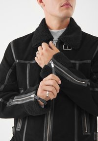Giacca in suede nera con colletto in shearling, polsini con zip e hardware in argento. Caratterizzata da un design strutturato con texture a contrasto.