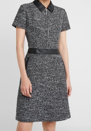 Kobieta ubrana w czarno-białą teksturowaną sukienkę o krótkich rękawach, sięgającą do kolan, z czarnym skórzanym kołnierzem, paskiem w talii i suwakiem z przodu.