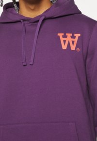Fialová mikina s klokaní kapsou vpředu, na levé části hrudi zdobena korálově zbarveným logem. Měkká tkanina s ribovanými manžetami a nastavitelnou kapucí se šňůrkou.