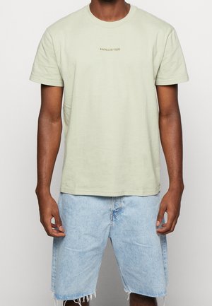Lysegrøn T-shirt med korte ærmer og "HOLLISTER" præget i en beige tone, kombineret med lyseblå denimshorts med frynser.