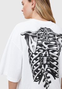T-shirt bianco oversize con una grande grafica nera che rappresenta una gabbia toracica e orecchie di cane sul retro. Materiale di cotone morbido con maniche corte.