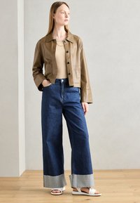 Veste en cuir marron taille cropped avec boutons noirs, associée à un jean bleu à jambes larges avec ourlets effilochés et des sandales blanches.