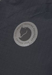 Fjällräven-Emblem auf strukturiertem, dunklem Stoff mit einem Rautenmuster. Runder grauer Patch zeigt eine stilisierte Outline eines Fuchses und Text.