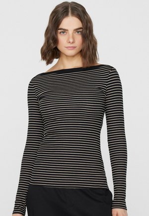 Junge Frau mit schulterlangem braunem Haar, die einen schwarz-beigen, horizontal gestreiften Langarmoberteil und schwarze Hosen trägt und nach vorne schaut.