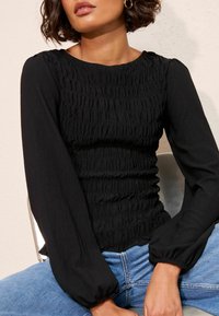 Czarna bluzka z długimi, bufkami oraz fakturowanym detalem marszczenia. Dopasowana góra z prążkowanej tkaniny, zestawiona z jasnoniebieskimi dżinsami.