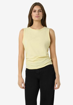 Žena s rovnými hnedými vlasmi, oblečená v bezrukávovom bledozlatom topu a čiernych nohaviciach, stojaca pred jednoduchým bielym pozadím.