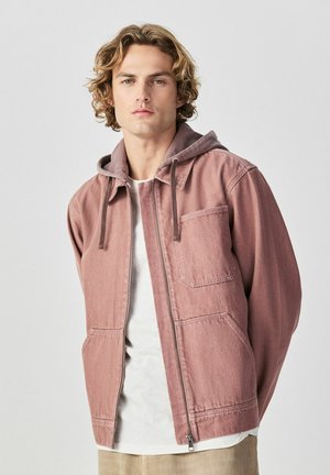 Mladý muž s kudrnatými blond vlasy, oblečený v prachově růžové bundě na zip přes bílé tričko, stojící před jednoduchým šedým pozadím.