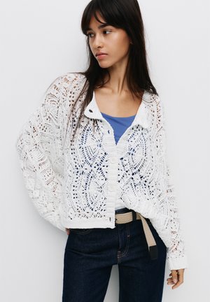 Cardigan bianco a maglia aperta con un motivo in pizzo geometrico, dotato di colletto e bottoni, indossato sopra una maglietta blu, abbinato a jeans scuri.
