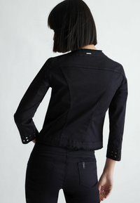 Musta farkkutakki, jossa on kolmannen osan hihat, ja jonka hihoissa on pitsileikkauksia sekä röpelöinen helma. Takakatsaus näyttää muotoiltua suunnittelua.