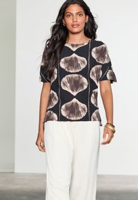 Black shirt with beige shell pattern, short sleeves, and a straight cut, paired with light beige loose pants. Smooth fabric texture.