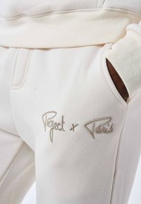 Pantaloni della tuta color crema in morbido tessuto, con vita a coste e logo ricamato "Project x Paris" in beige nella zona della tasca.