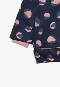 Tmavě modrá prošívaná bunda s růžovými a modrými kruhovými vzory, elastickými manžetami a zlatým lemem podél spodního okraje. Hladká, lesklá textilie.