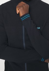 Svart långärmad zip-up tröja med texturerad tyg och blåa accentlinjer på muddar. Med hög krage och åtsittande design.