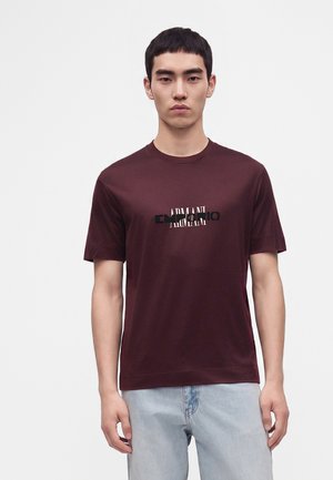 Jeune homme portant un T-shirt bordeaux avec un texte de logo Armani superposé en blanc et noir, associé à un jean bleu clair, debout devant un fond uni.