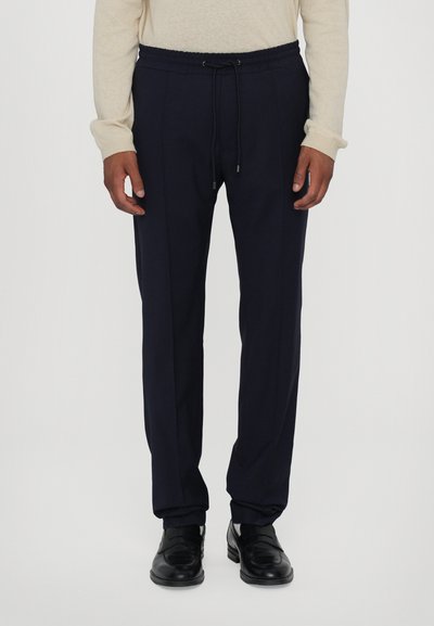 Navy blue sweatpants with a drawstring waistband, tapered leg design, and side pockets, paired with black shoes and a light sweater.