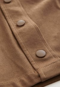 Bruine suede stof met een rij metallic drukknoopjes, zichtbaar stiksel en een gladde textuur, met de nadruk op het knoopdetail.