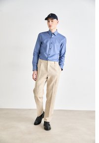 Light blue fitted shirt with black buttons, paired with beige textured trousers and black shoes. Additionally, a navy cap with logo is worn.