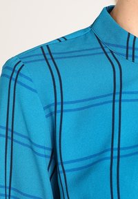 Maglietta turchese con colletto, caratterizzata da righe orizzontali e verticali blu navy, texture liscia e design di vestibilità standard.