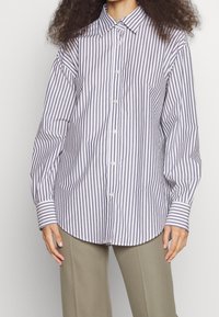 Persona che indossa una camicia bianca a maniche lunghe con strisce nere verticali e pantaloni beige, in piedi davanti a uno sfondo chiaro.
