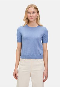 Maglione azzurro chiaro a maniche corte, realizzato in un tessuto testurizzato e lucido. Presenta un collo rotondo e un orlo a coste, abbinato a pantaloni color crema.