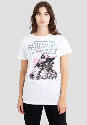 Young woman wearing a white Star Wars t-shirt with a graphic of Darth Vader and characters in black and pink, standing against a plain background.